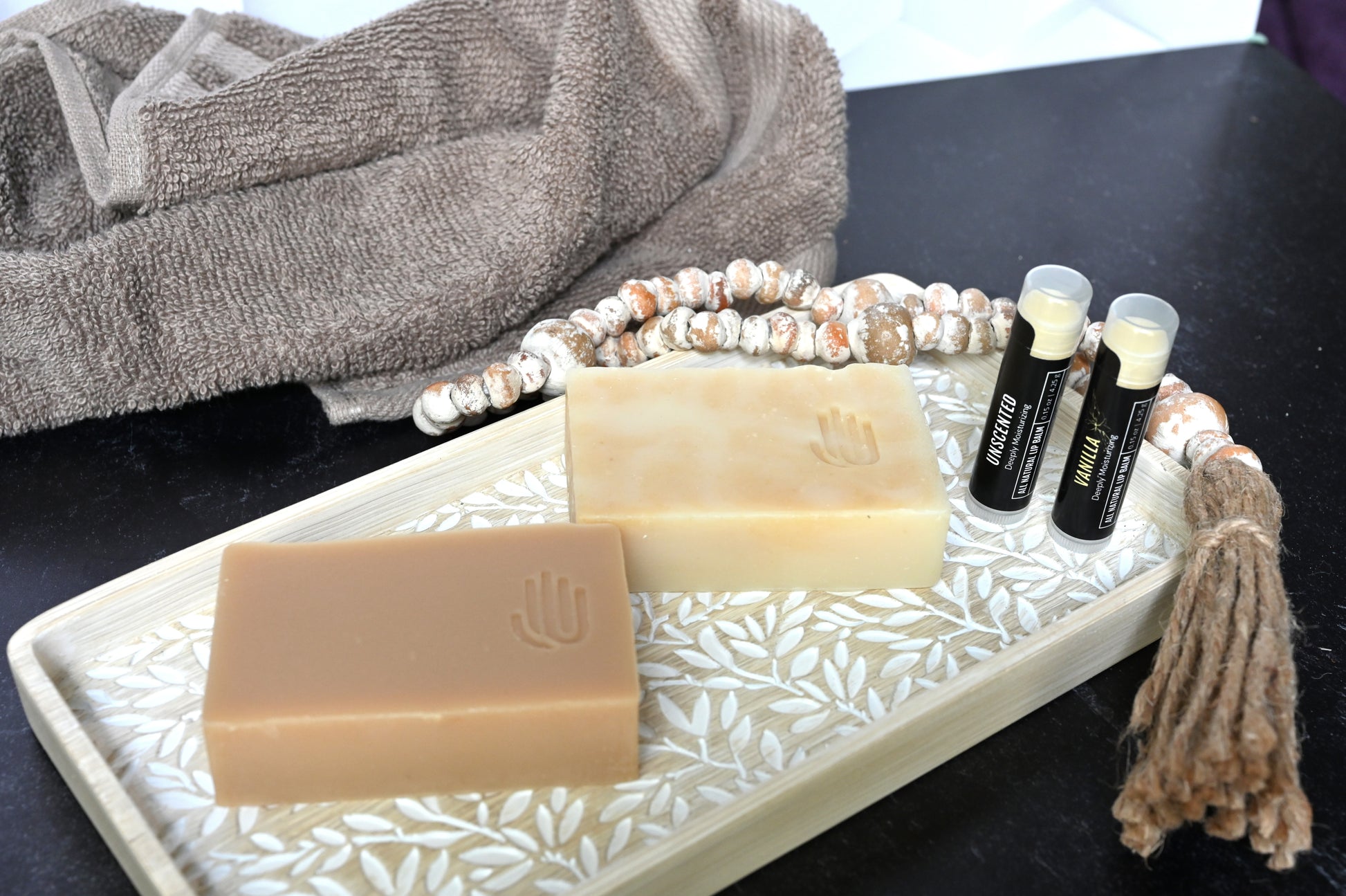 2 all natural lip balms with black labels sitting upright on a decorative wooden tray next to 2 taupe colored soaps with a tan towel in the background sitting on a black bathroom sink