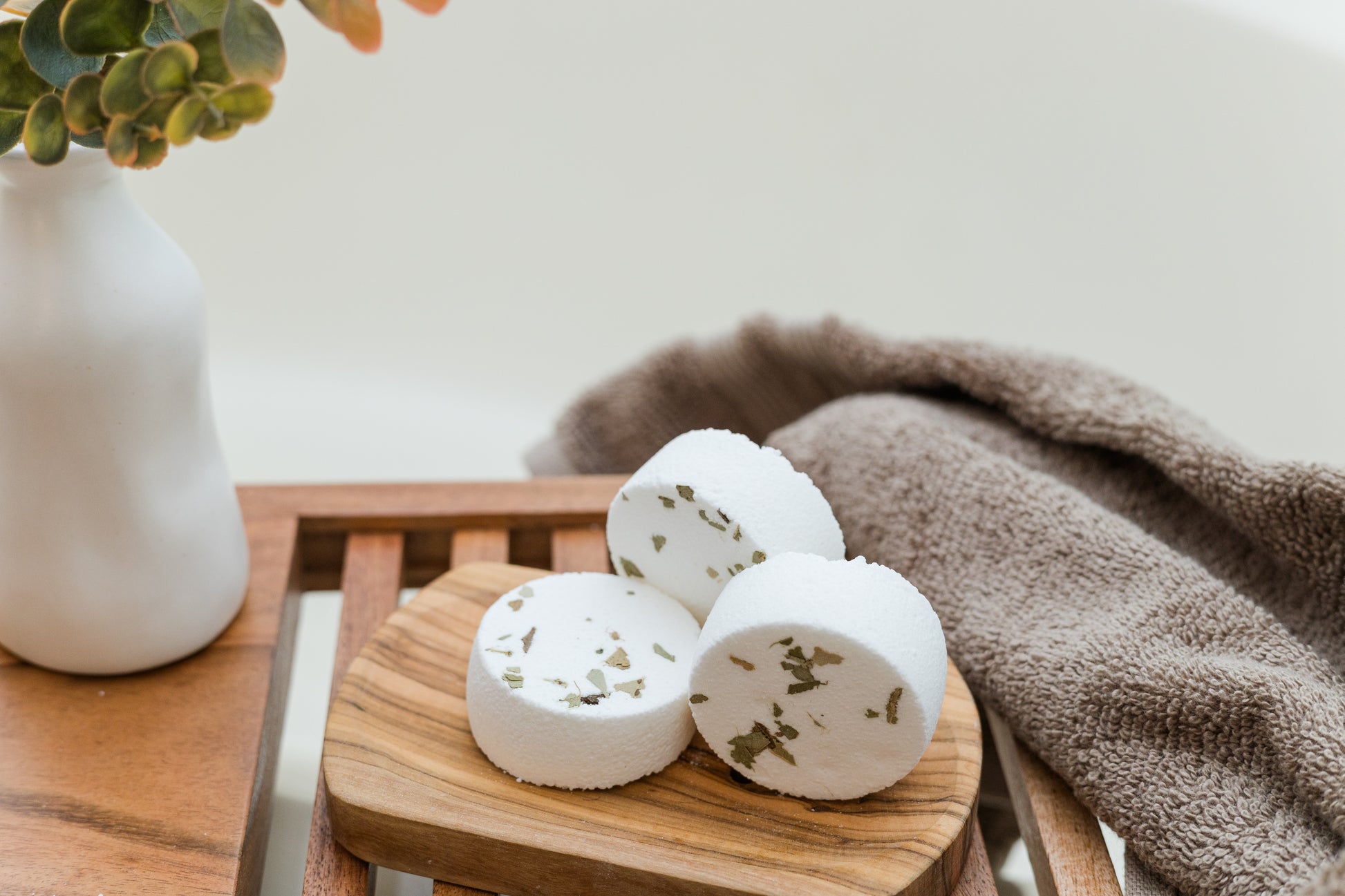 three eucalyptus shower steamers on a wooden dish with a vase and eucalyptus sprigs behind it