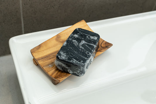 wooden soap dish that curves up on both sides with black soap sitting on it placed on a bathroom sink